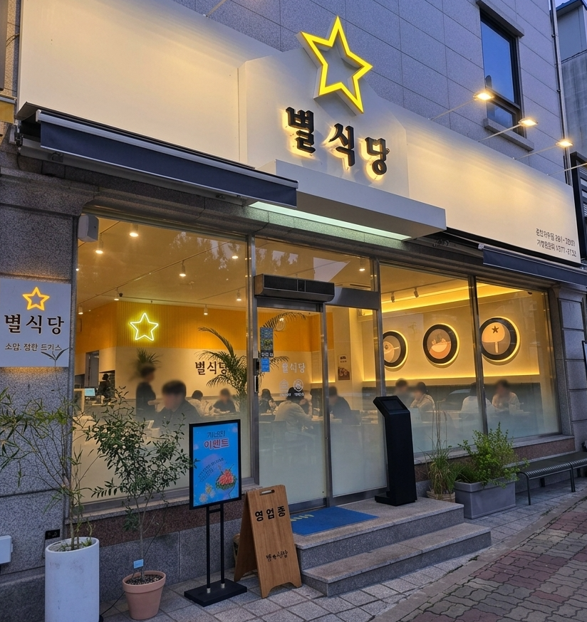 해질 무렵 춘천 시내, 깔끔한 흰색 외벽의 아담한 일식당 외관을 정면에서 살짝 비스듬히 잡은 컷. 통유리창 너머로 손님들이 앉아 있는 실루엣과