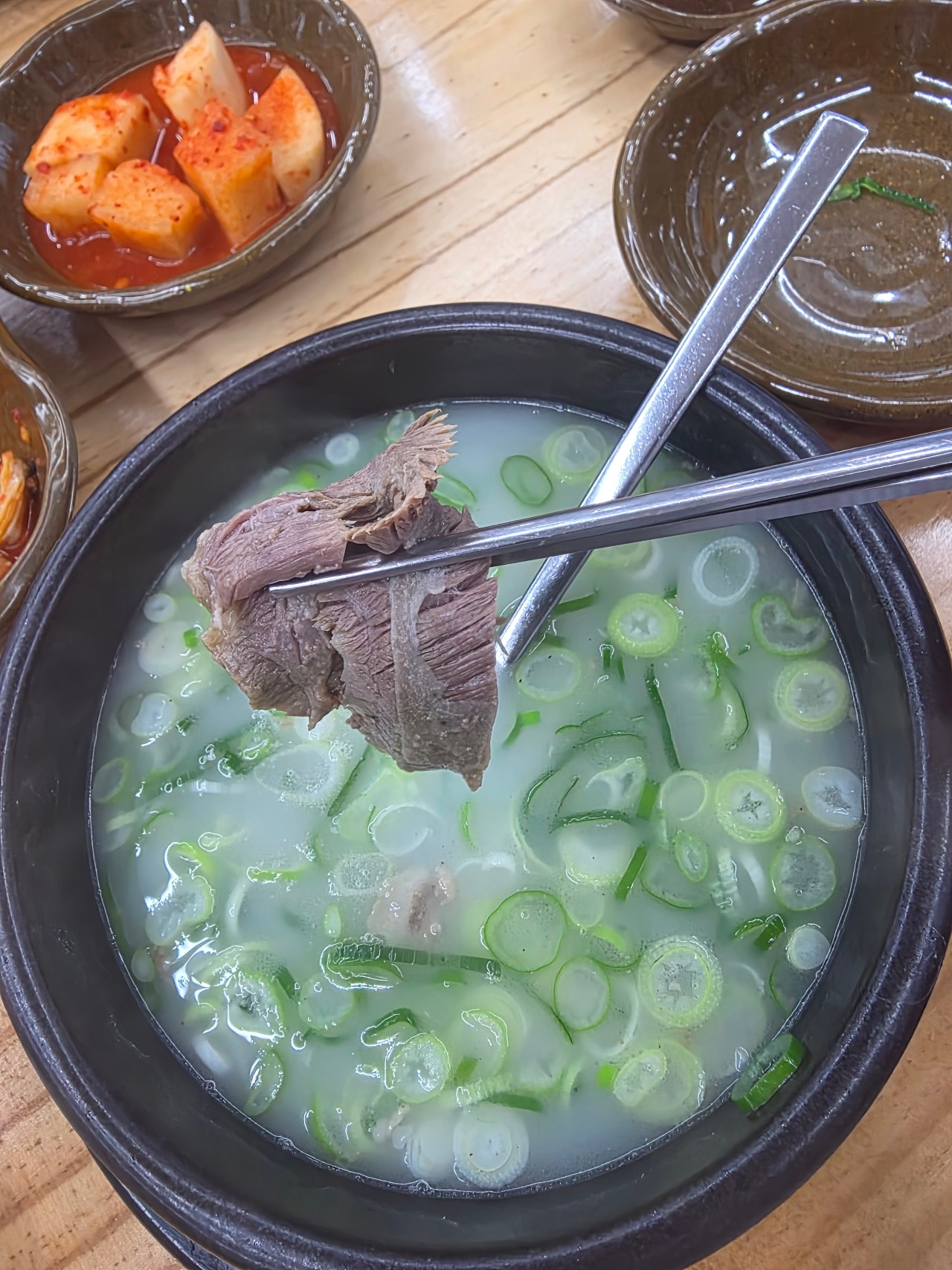 이른 아침 국밥집 나무 식탁에서 젓가락으로 건져 올린 설렁탕 수육 한 점. 결이 갈라진 단면에서 뽀얀 국물이 떨어지기 직전이고, 뒤로는 검은 뚝배기와 붉은 깍두기 종지가 놓여 있다